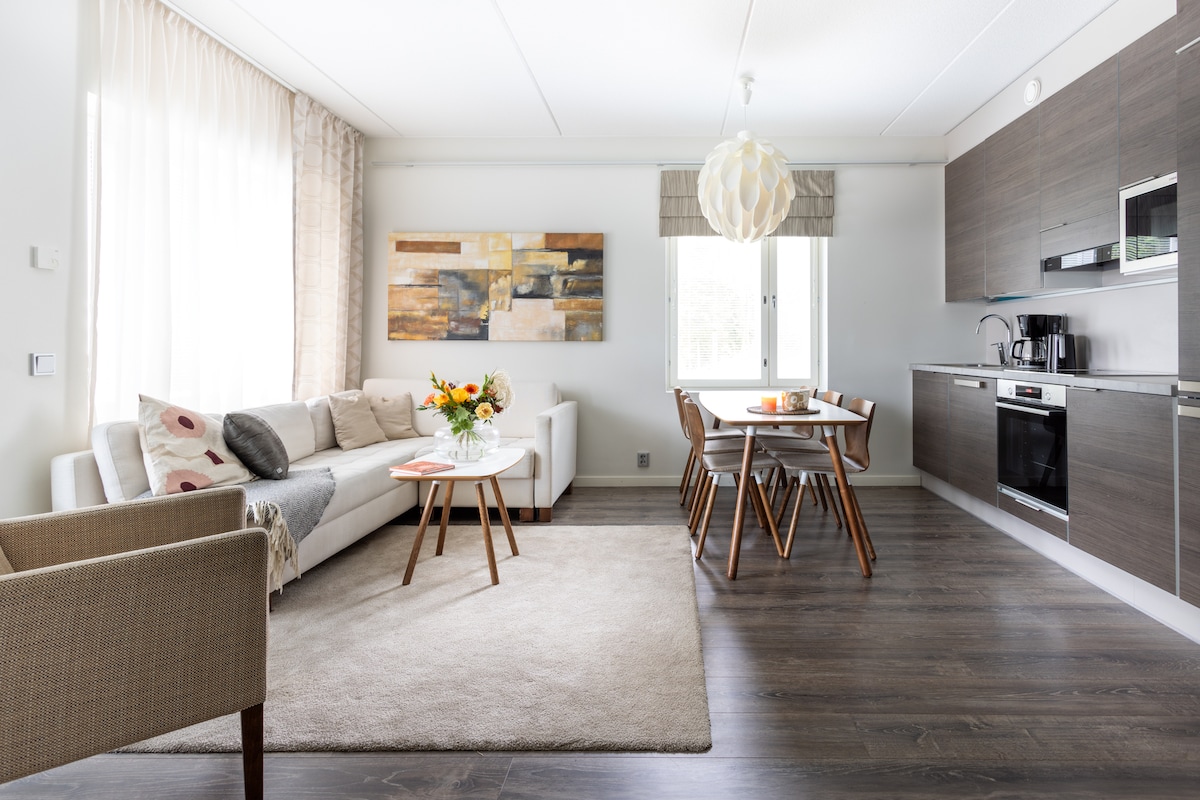A modern living area features a light-colored sofa adorned with patterned cushions and a textured throw. A round table with wooden legs is surrounded by four chairs. A bouquet of flowers rests on the coffee table, while a large window provides natural light.