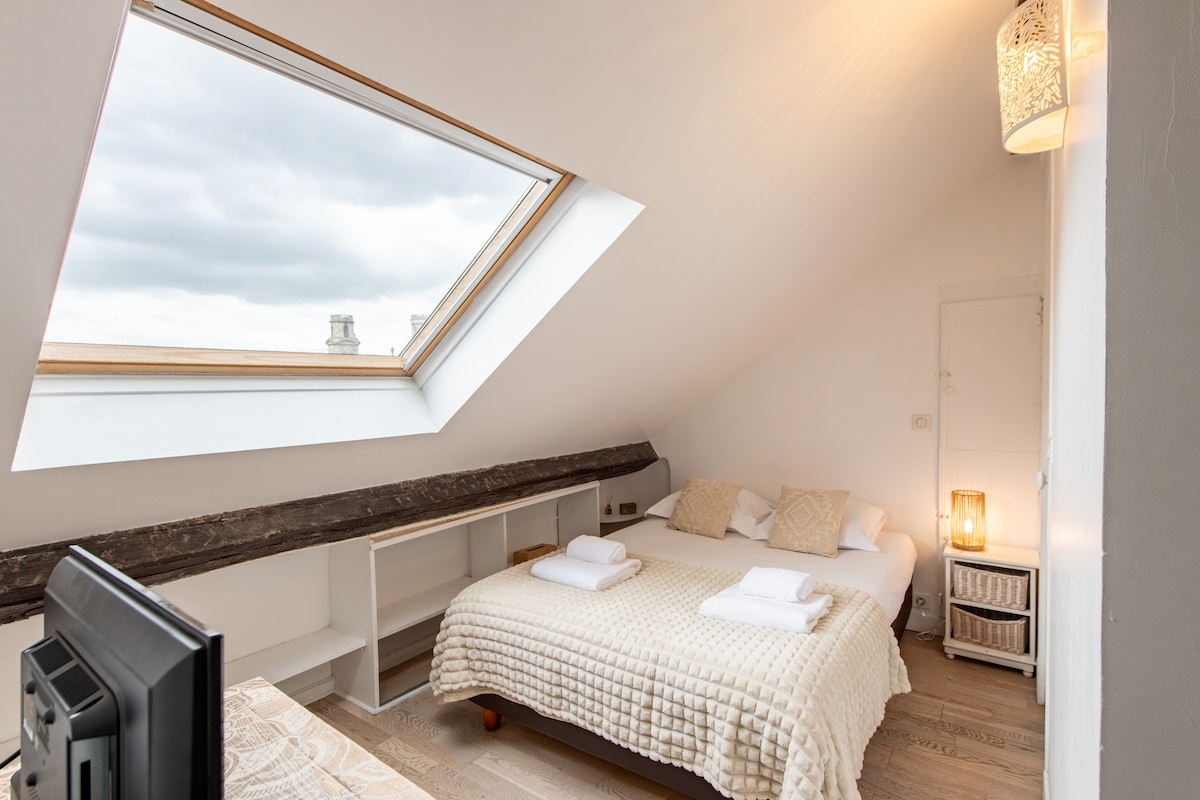 A spacious bedroom is featured with a comfortable double bed dressed in a light quilt. A large skylight allows natural light to illuminate the room. Shelving and a lamp provide functional and warm touches to the space, enhancing its cozy atmosphere.