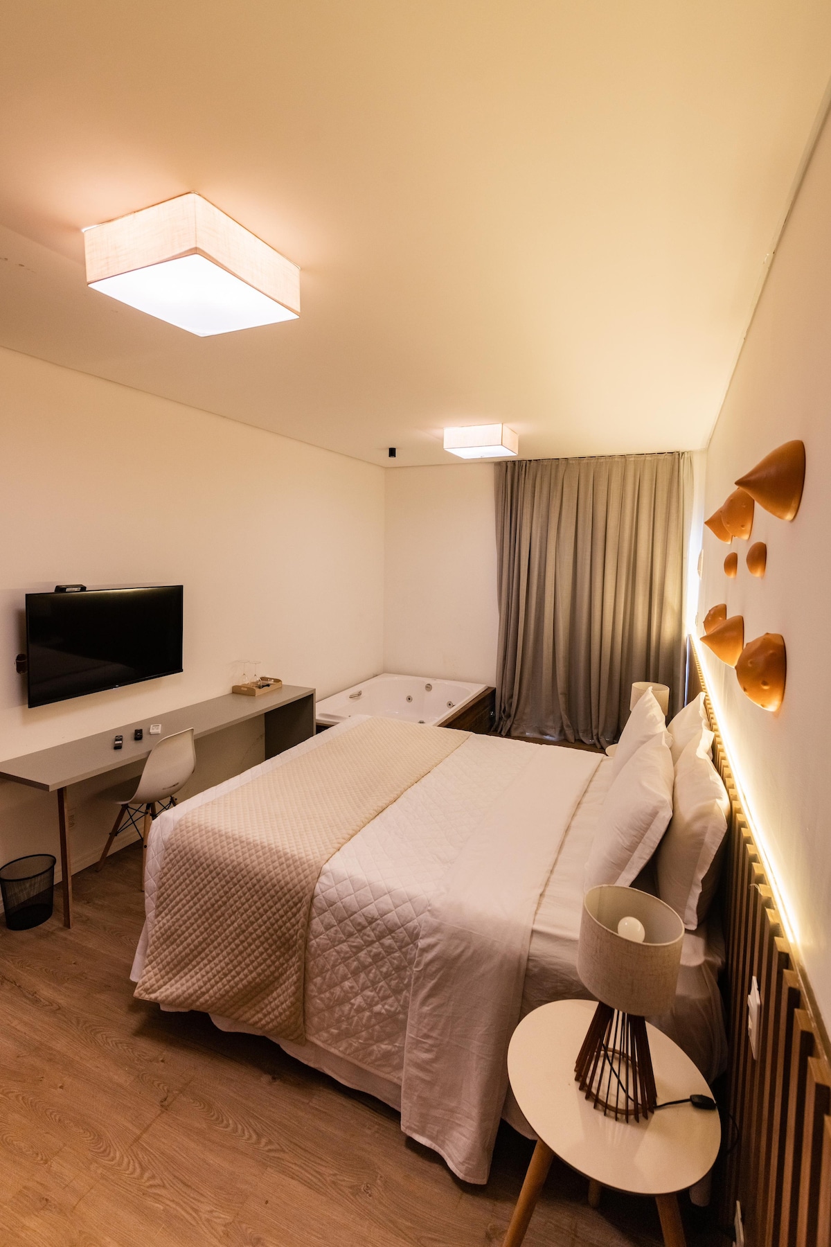 The room features a spacious bed dressed in light bedding, complemented by decorative wall accents. A flat-screen television is mounted opposite the bed, while a stylish desk and chair are positioned nearby. Soft lighting creates a calm atmosphere, and a bathtub is situated to one side.