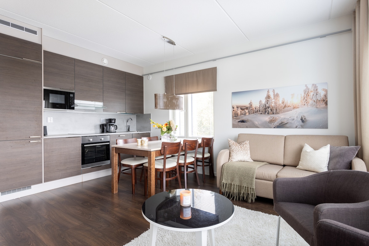 A modern living area is showcased, featuring a comfortable sofa and a dark armchair positioned beside a round coffee table. A dining table with wooden chairs is set against the kitchen, which includes sleek cabinetry and appliances. Natural light enters through a large window, illuminating the space.