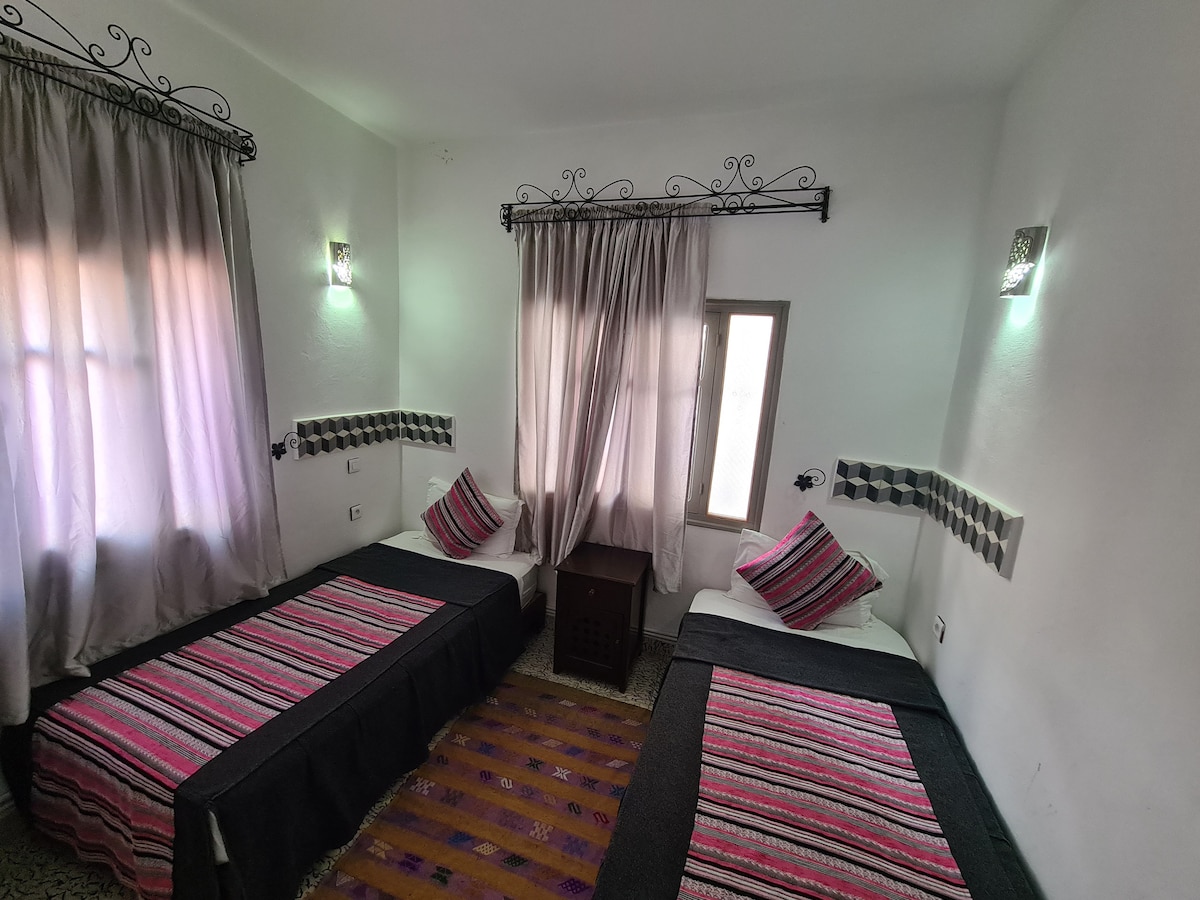 A well-lit room features two twin beds dressed in dark blankets and colorful striped accent pillows. Natural light enters through a window, which is framed by light curtains. Decorative wall sconces provide soft lighting, while a patterned area rug adds warmth to the space.