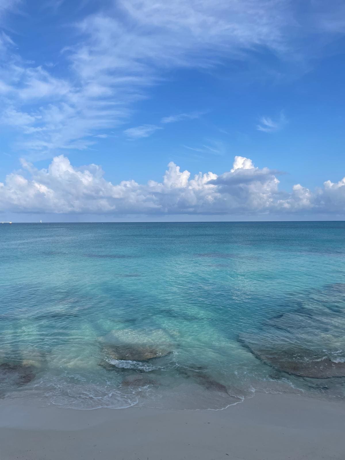 A serene ocean view is presented, featuring calm, turquoise waters gently lapping at a sandy shore. The sky is adorned with soft, white clouds and deep blue tones. Subtle rock formations can be seen beneath the water's surface, adding texture to the scene.