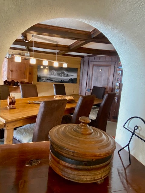 A rustic dining area is framed by an arched entryway. A long wooden table is surrounded by dark upholstered chairs, and warm lighting is provided by hanging pendant lights. A decorative wooden bowl sits prominently on a nearby surface, adding an inviting touch to the space.