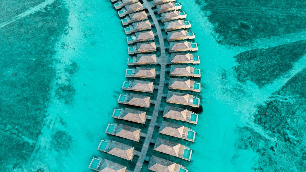 An aerial view showcases a series of water villas connected by wooden pathways, set against clear turquoise waters. The structured layout highlights the villas' thatched roofs and surrounding natural features, emphasizing the serene beauty of the resort's setting.