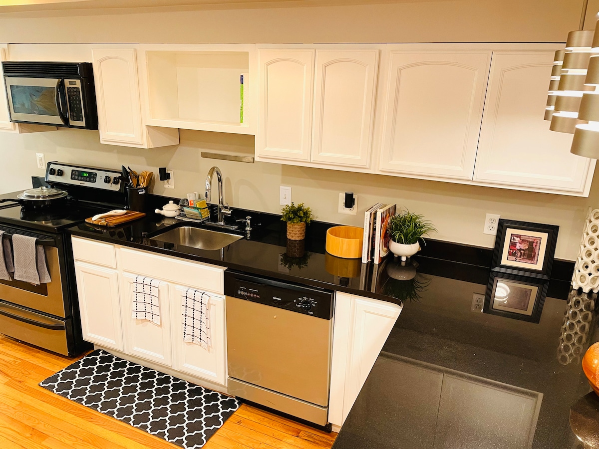 A modern kitchen features sleek black countertops and white cabinetry. Stainless steel appliances include a refrigerator and dishwasher. An organized sink area is complemented by decorative plants and kitchen towels. A patterned mat lies beneath the prep area, enhancing the functional layout.