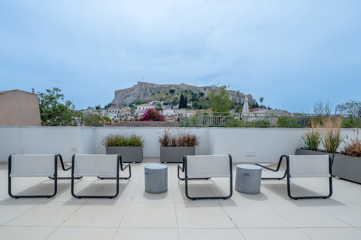 A spacious shared terrace is presented, featuring modern seating arrangements consisting of six minimalist chairs and one round table. Lush greenery is seen in planter boxes along the terrace edge, with a view of the Acropolis visible in the background against a cloudy sky.
