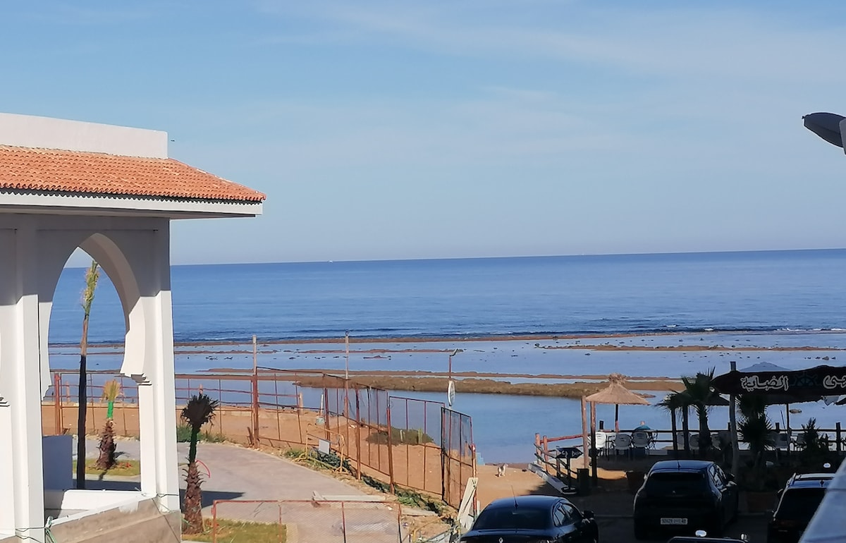 A serene view over a calm sea stretches towards the horizon, with gentle waves visible. The near shoreline features sandy areas bordered by palm trees and outdoor seating. A building with an archway stands in the foreground, contributing to the coastal ambiance.