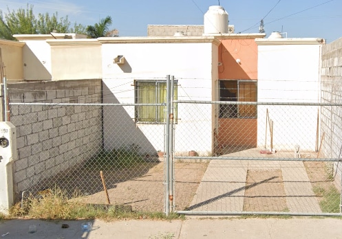 The exterior of a single-story home is presented, featuring a fenced yard with a mix of gravel and grass. A small path leads to the entrance, where a door is framed by two windows with beige curtains. The walls exhibit a light color scheme with a contrasting orange section.