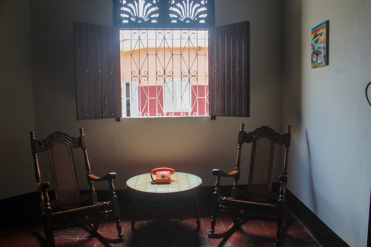 Two traditional rocking chairs are positioned beside a small round table, which holds a vintage telephone. A large window adorned with decorative ironwork allows natural light to fill the space, providing a view of the outside. A small colorful painting is mounted on the wall.