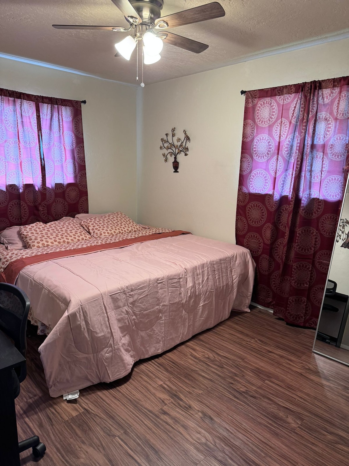 The bedroom features a spacious bed covered with a soft pink blanket, complemented by patterned pillows. Natural light filters through sheer curtains in a deep red hue. A mirror and a small desk are present, adding functionality to the space, which is finished with warm wood flooring.