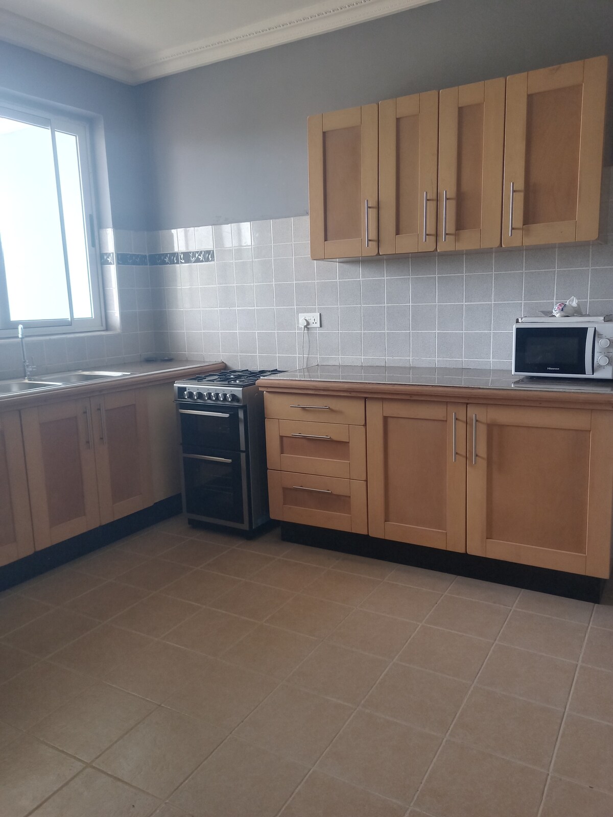 A modern kitchen is designed with wooden cabinetry, showcasing ample storage space. Stainless steel appliances, including a stovetop and microwave, are integrated into the layout. A window allows natural light to fill the room, illuminating the tiled backsplash and countertops.