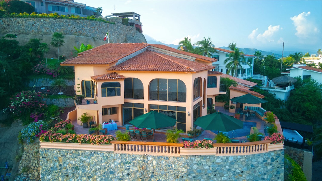 The exterior of the house is showcased with a terracotta roof and large arched windows. Surrounding greenery enhances the view, while shaded patios and colorful flower beds create a serene outdoor space. The location on a hillside allows for expansive views of the landscape.