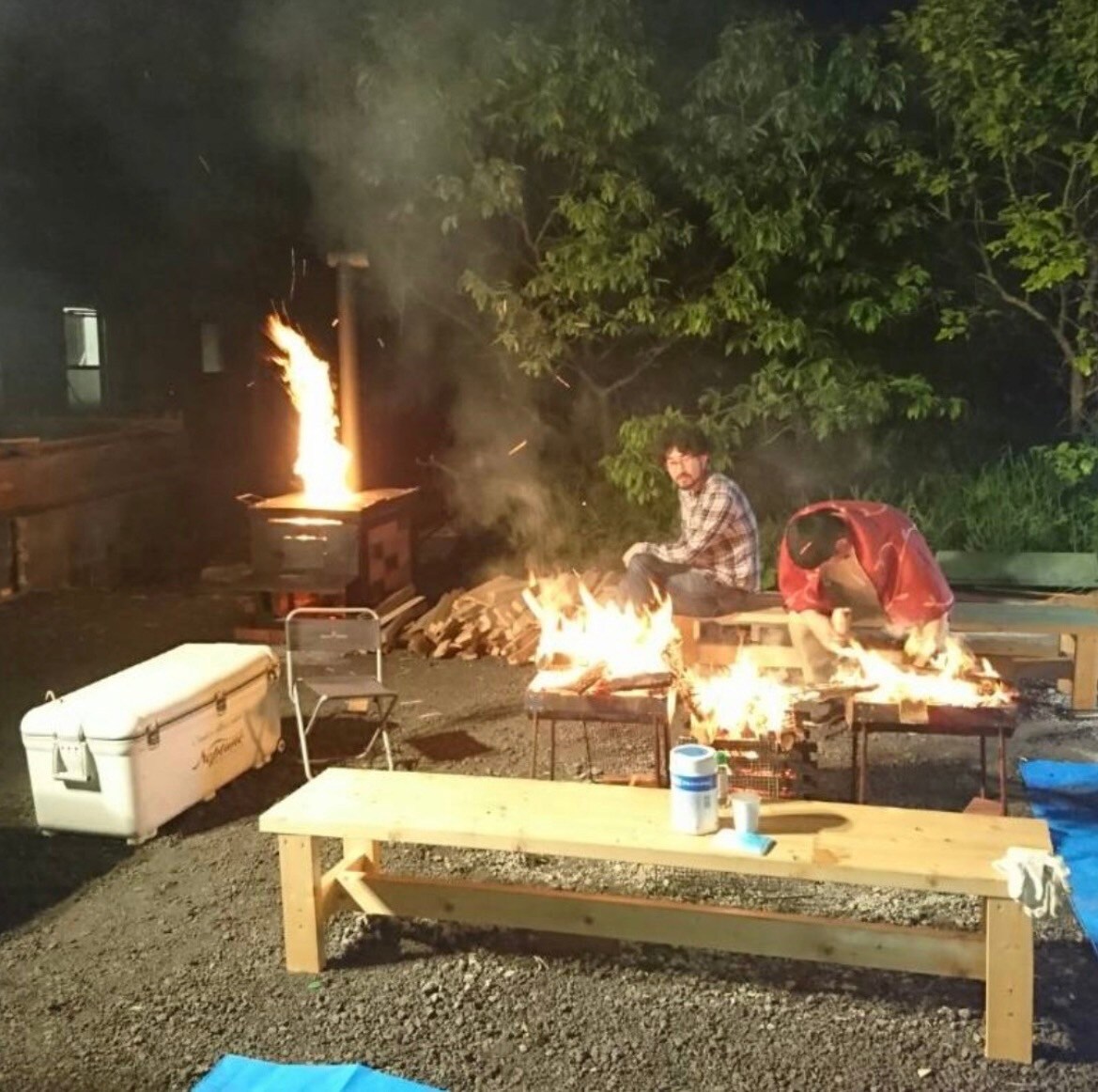 A nighttime outdoor setting features a fire pit with flames, surrounded by two individuals preparing food. Wooden benches are positioned nearby for seating. A cooler and a stack of wooden logs are also visible, contributing to the communal cooking experience.