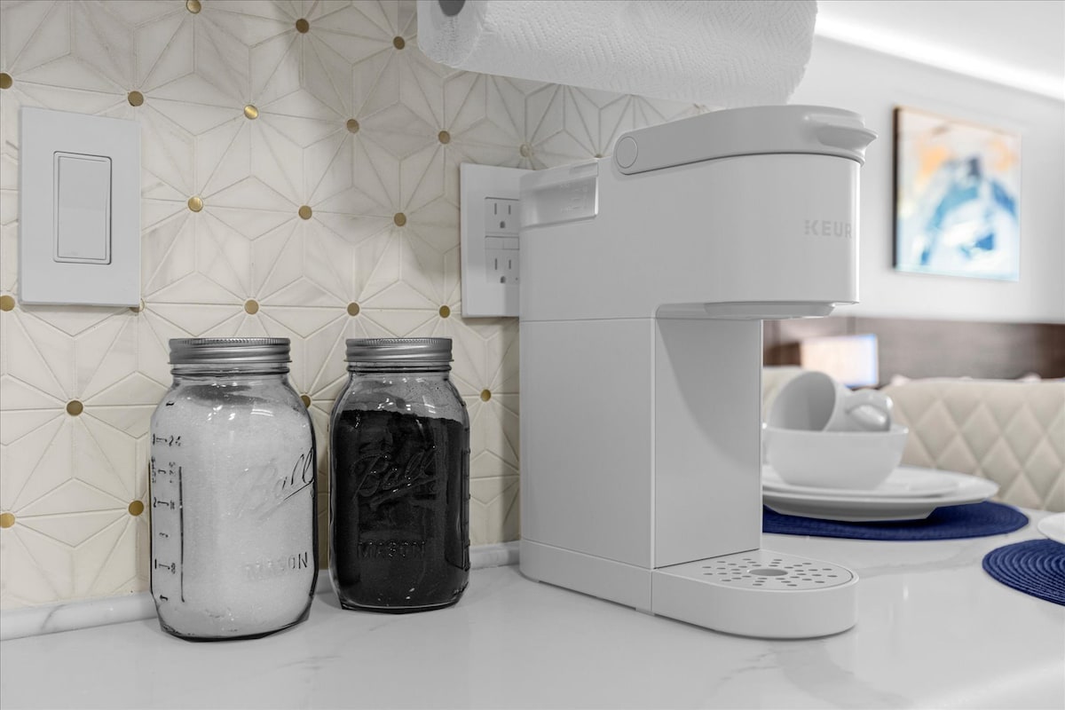 A modern coffee station is displayed, featuring a white Keurig coffee machine alongside two glass jars filled with sugar and coffee. The backdrop showcases a geometric tile design, adding a stylish touch to the kitchen area.