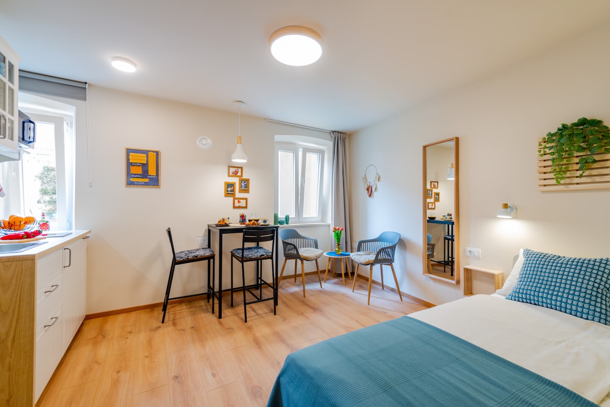 A well-lit studio apartment features a spacious bed with a light blue bedspread. A small dining table with high chairs occupies one corner, while a cozy seating area includes two comfortable chairs. Natural light streams through the window, complemented by wall decor and a mirror.