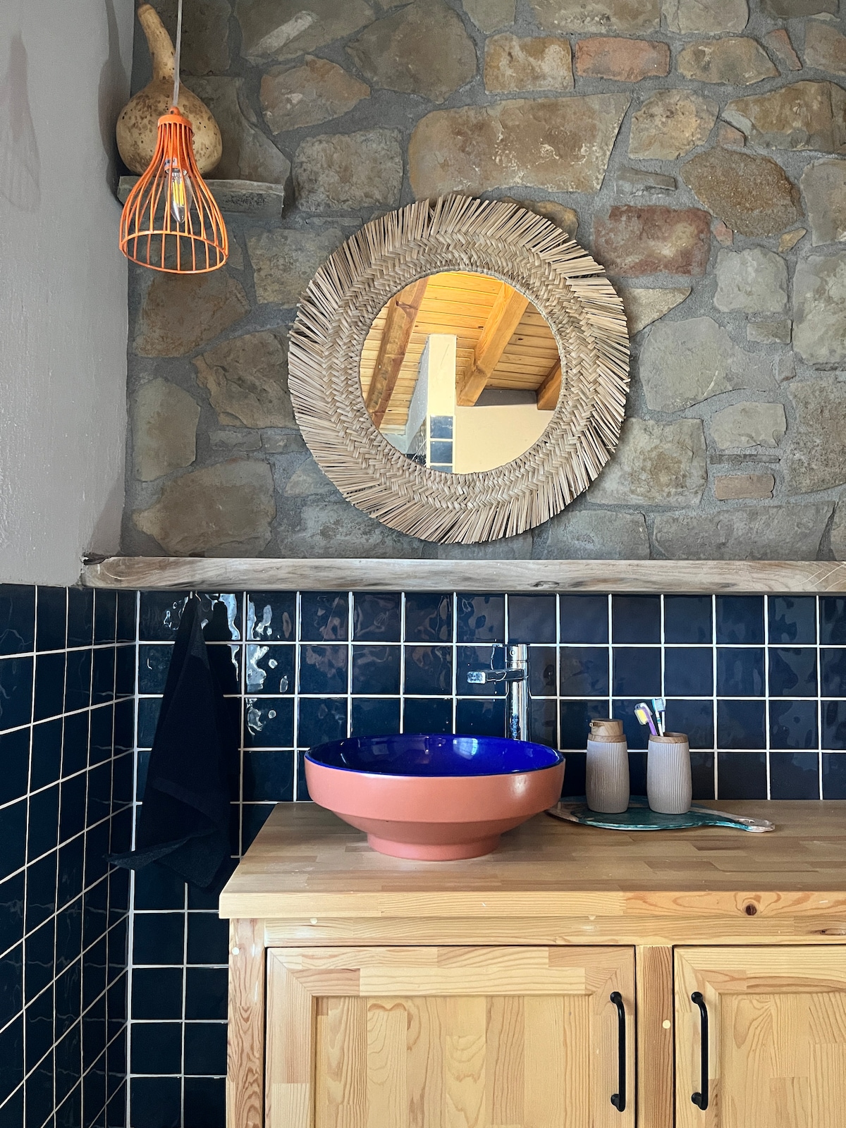 A stone wall serves as the backdrop for a wooden countertop with a large, colorful bowl. A round mirror with a natural frame hangs above, while a small shelf holds several glass and ceramic containers. An orange light fixture provides illumination.