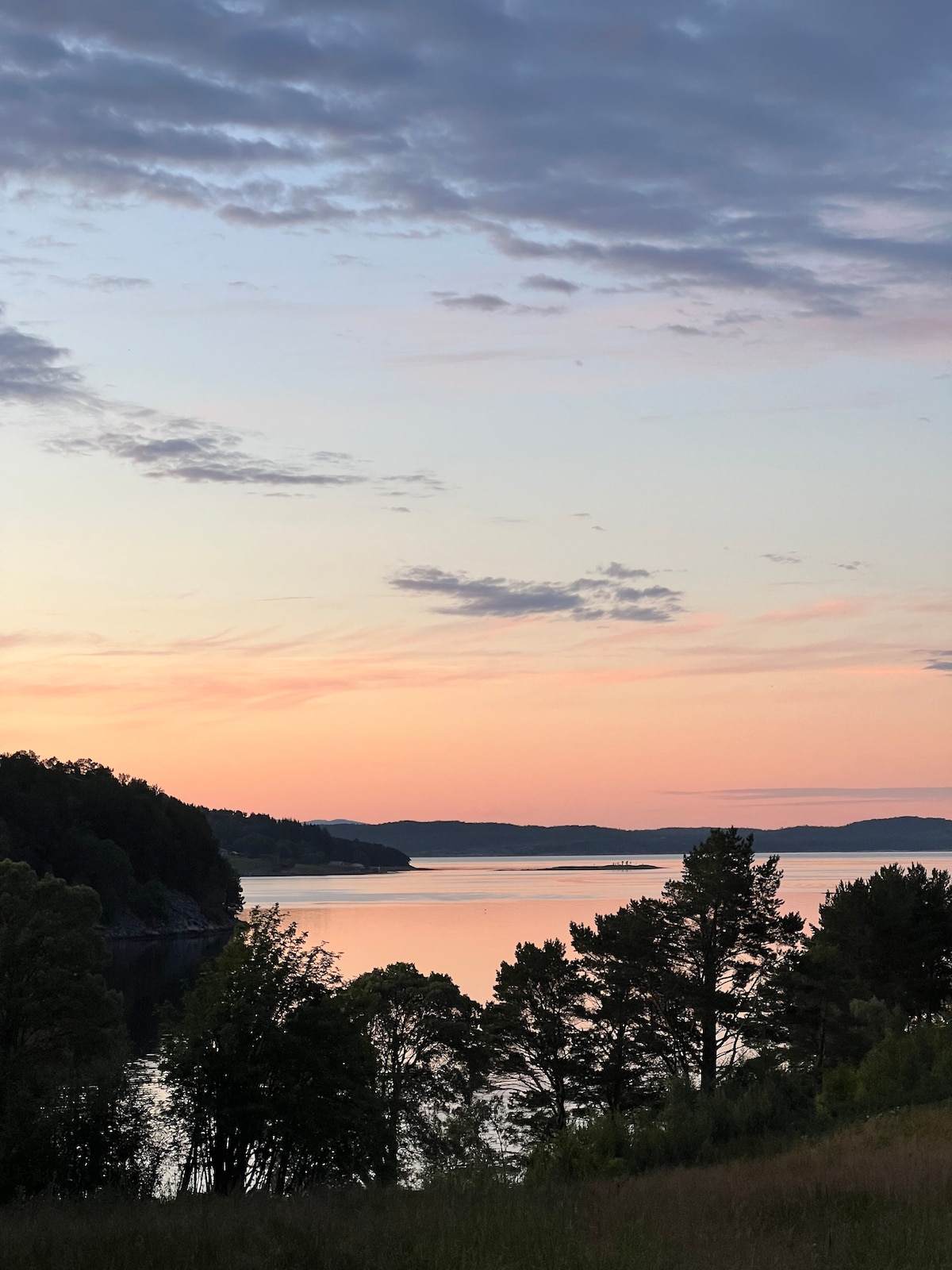 A serene lakeside view features soft pastel hues in the sky as the sun sets, reflecting on the calm water. Silhouette of trees frames the scene, while the landscape gently slopes towards the lake, inviting a sense of tranquility.