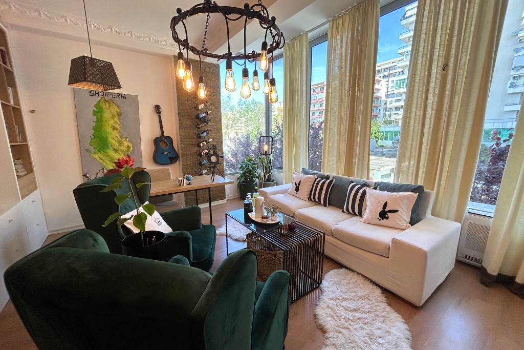 A spacious living area features large windows allowing ample natural light. A white sofa and two green armchairs surround a glass-topped coffee table. Decorative plants, a guitar, and wall art enhance the ambiance. Sheer curtains soften the sunlight, creating a bright yet comfortable space.