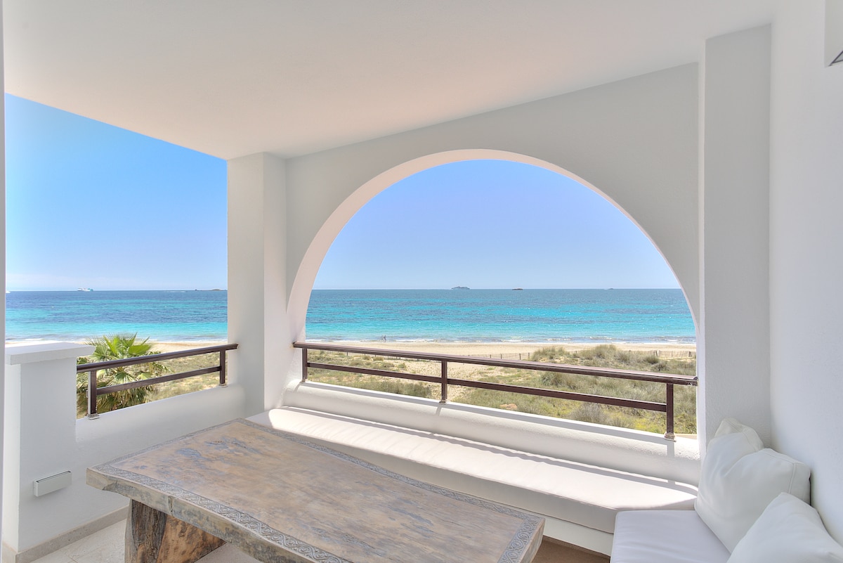 An open terrace features a large wooden table with a built-in seating area, inviting relaxation. The view showcases a vibrant blue sea meeting a clear sky, framed by a large arched opening. Lush greenery is visible in the foreground, enhancing the natural setting.