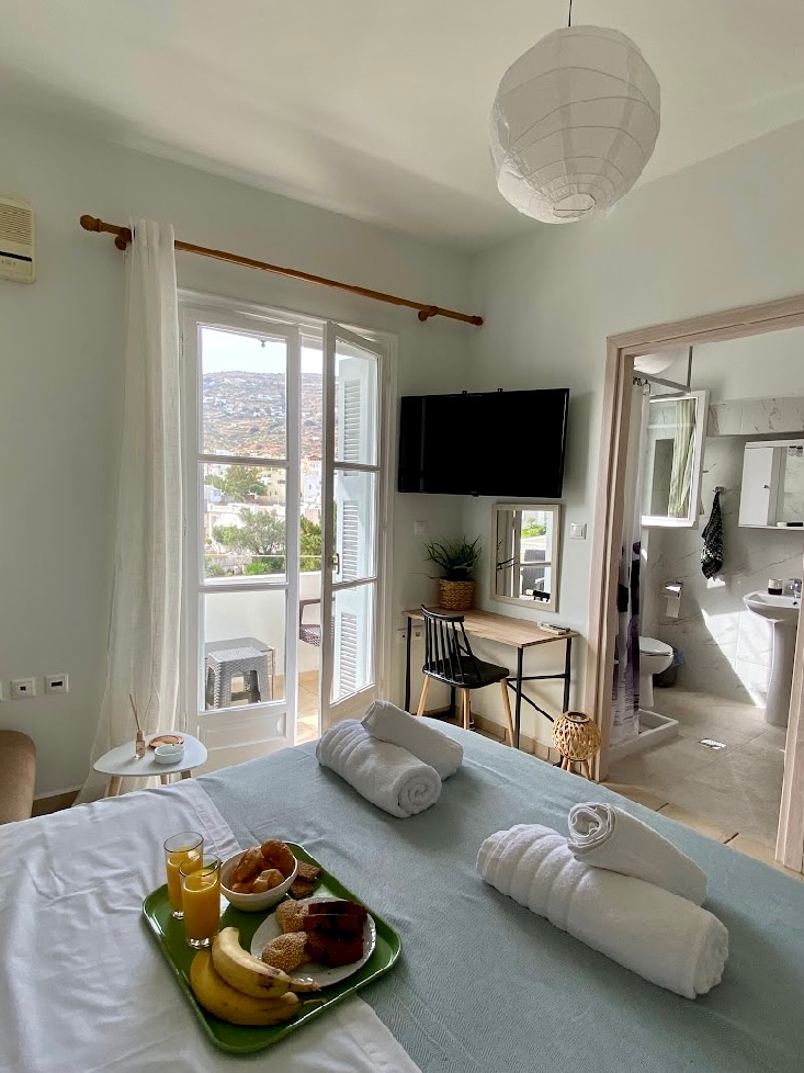 A well-lit studio is depicted with a view outside through the double doors, showcasing a serene landscape. A cozy bed sits in the foreground, complemented by neatly arranged towels. A small table with breakfast items and a work desk is positioned nearby, enhancing functional space.