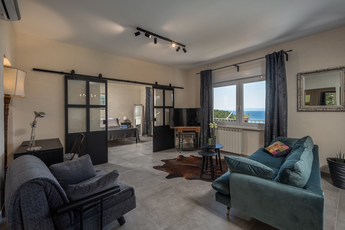 A spacious living area features modern furnishings, including a dark sofa and a light teal couch. Large windows provide natural light and views of the sea. A small table with a vase and a mirror add to the room's decor, while sliding doors separate the spaces.