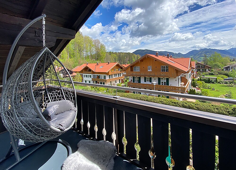 A private balcony features a hanging chair, providing a comfortable spot to enjoy views of the surrounding alpine landscape and neighboring houses. Lush greenery and distant mountains create a serene backdrop, with ample natural light illuminating the space.