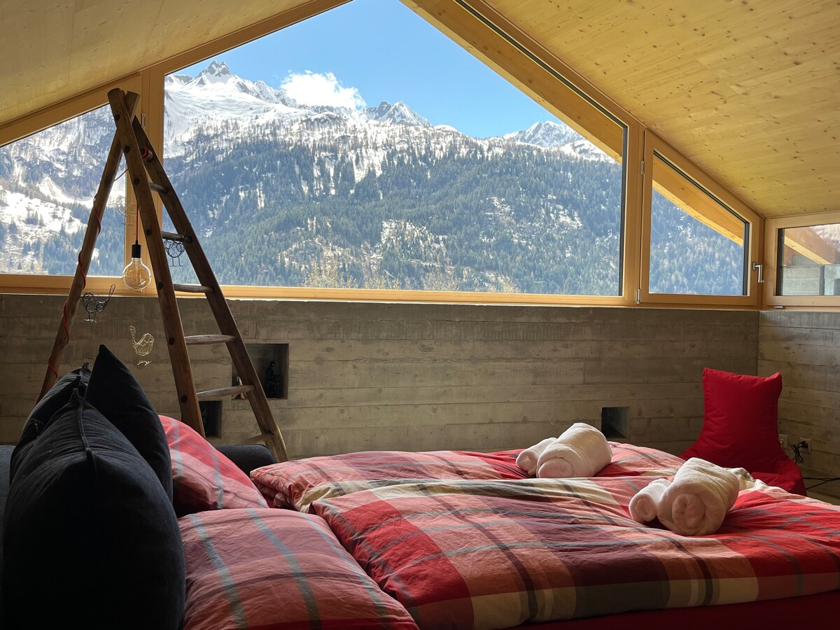 A spacious sleeping area features large windows that frame majestic mountain views. Two beds are adorned with plaid blankets, complemented by soft towels. A wooden ladder adds a rustic touch, while a cozy red cushion invites relaxation.