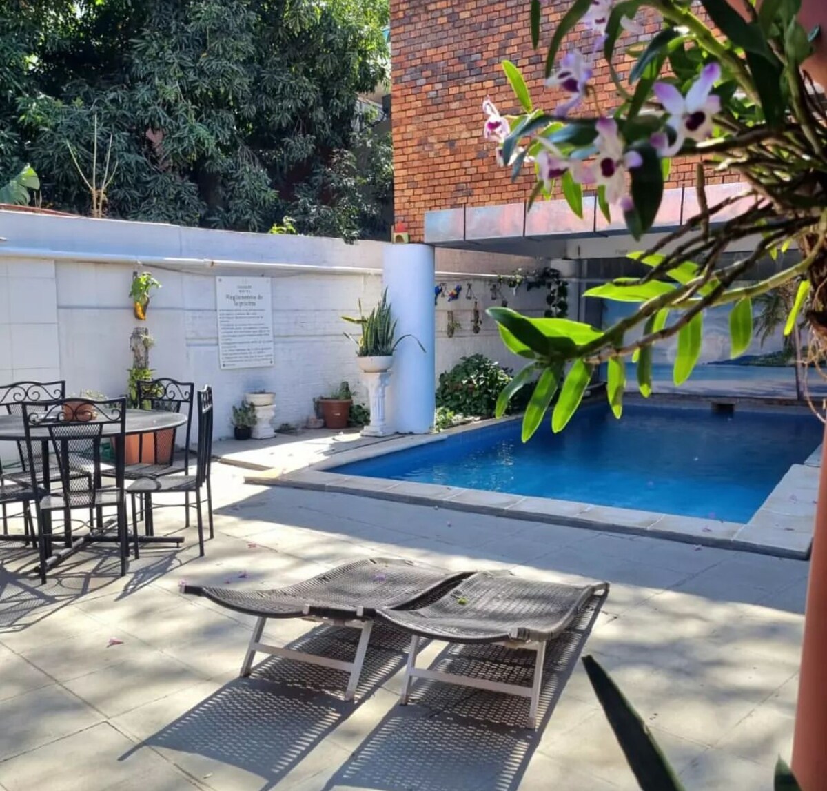An outdoor leisure area features a swimming pool surrounded by lush greenery. Patio furniture, including a dining table with chairs, is arranged on a paved surface. Two lounge chairs are positioned at the edge of the pool, inviting relaxation in a serene environment.