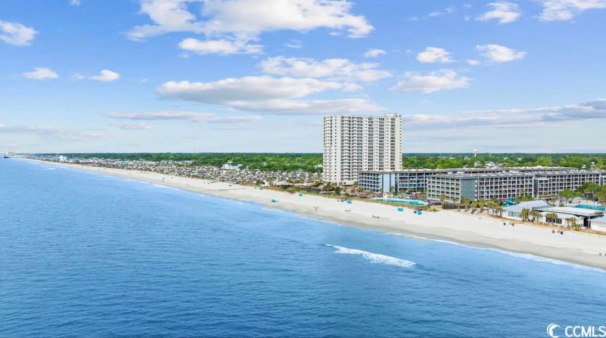 The image captures a scenic beach view, showcasing a wide stretch of sandy shore alongside a calm blue ocean. A tall building stands in the background, with a collection of smaller structures visible near the water's edge. The sky is bright with scattered clouds.