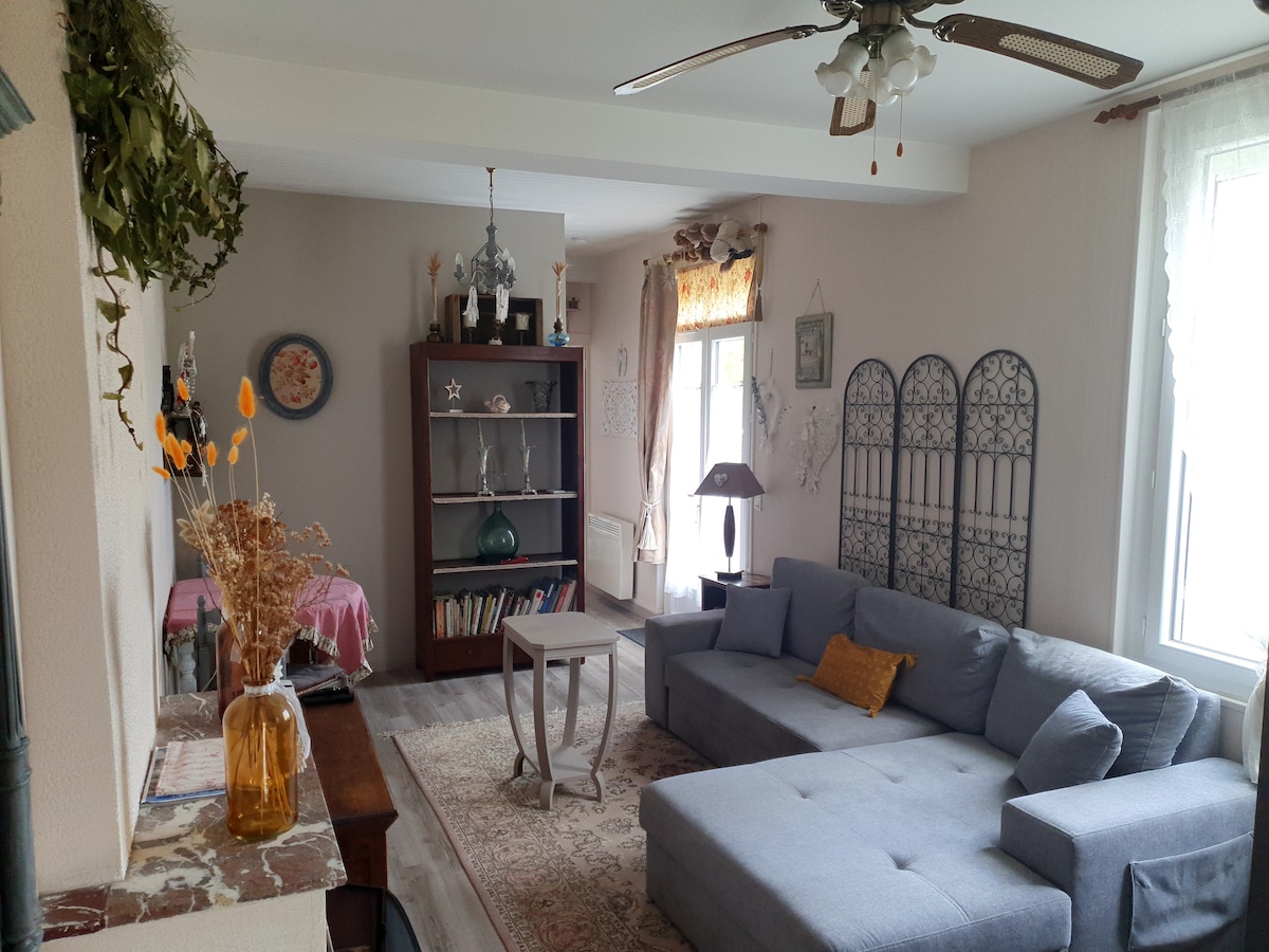 A spacious living area is showcased, featuring a comfortable gray sectional sofa and a small wooden table. A bookshelf holds various decorative items, and light filters through large windows adorned with sheer curtains, creating a warm and bright ambiance.
