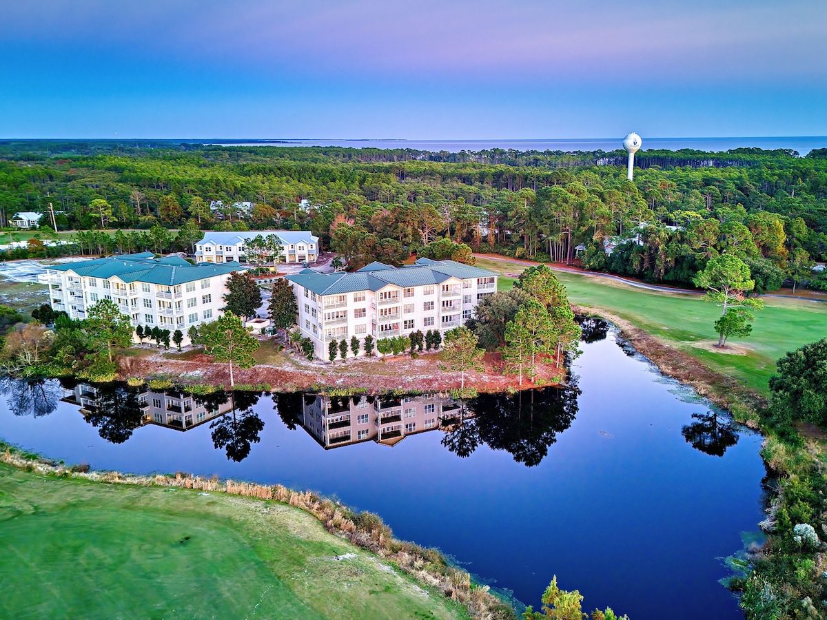 An aerial view captures the condo complex nestled alongside a serene lake, reflecting the surrounding greenery and blue sky. The golf course is visible in the foreground, emphasizing the tranquil setting and connection to nature.