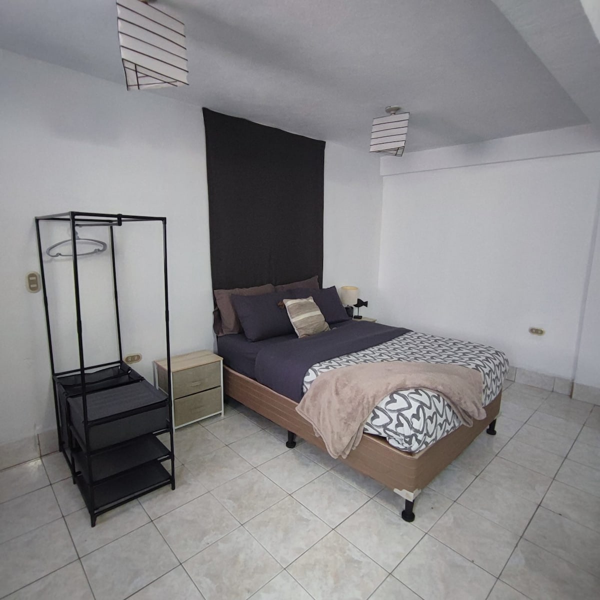 A spacious sleeping area features a comfortable bed dressed in neutral tones, accompanied by a nightstand with a lamp. A clothing rack stands nearby, and overhead lighting is provided by simple pendant lamps. The room is adorned with light-colored tiled flooring.