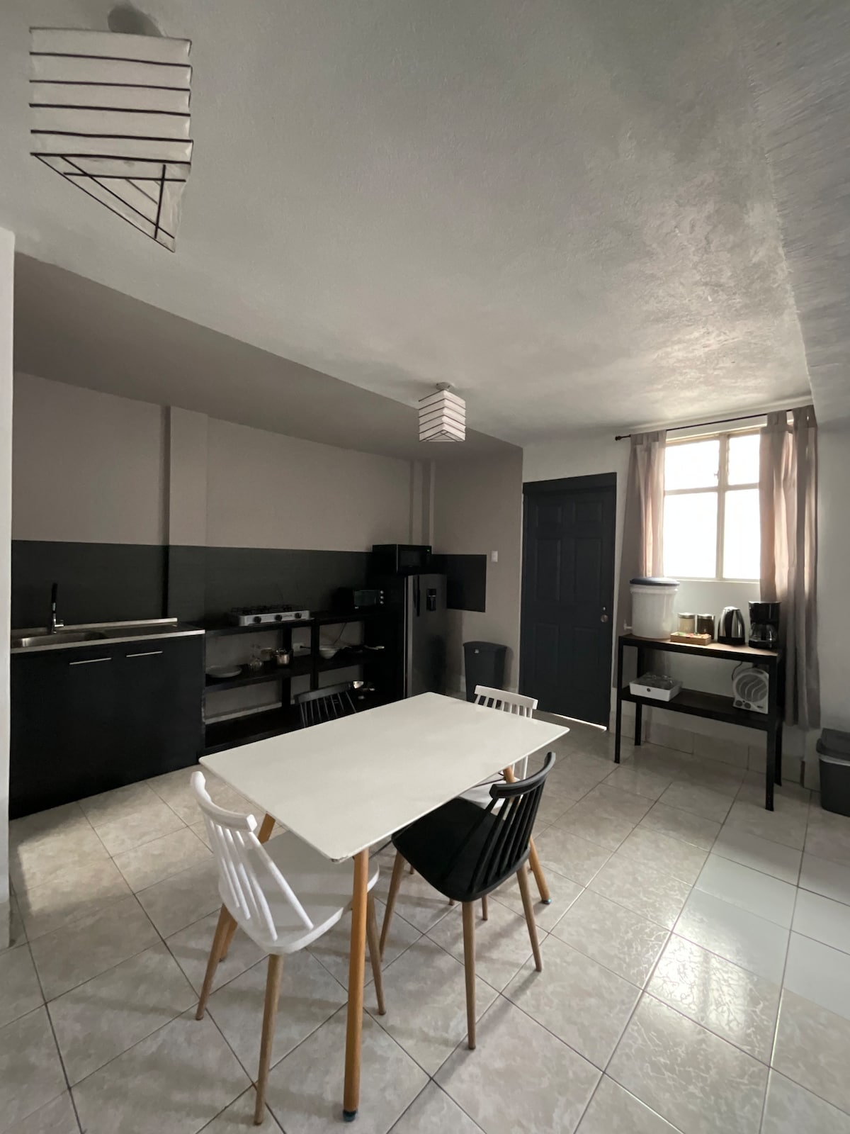 A modern kitchen area is shown, featuring a light-colored table surrounded by four chairs in varying designs. A dark cabinetry and appliances are visible along one wall, with natural light coming through a window draped with sheer curtains.