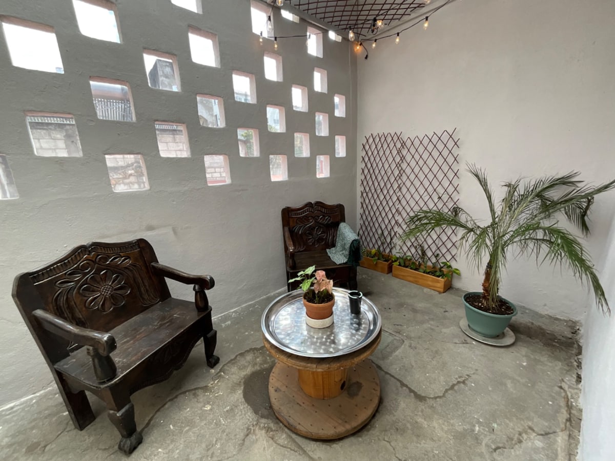 A private mini-patio features two dark wooden chairs and a round wooden table with a glass top. A small potted plant is centered on the table. A decorative trellis and a potted palm add greenery to the space, which is illuminated by soft string lights overhead.