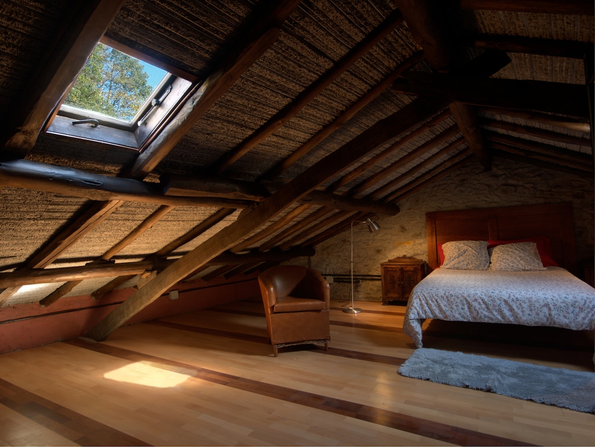 An attic bedroom is characterized by slanted wooden beams and a skylight allowing natural light to illuminate the space. A spacious bed is positioned beside a wooden nightstand, with a cozy armchair placed nearby, enhancing the room's charm.