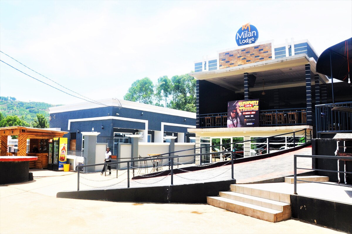 A modern building with a large sign reading 'Milan Lodge' is visible. The structure features an outdoor seating area with tables and chairs overlooking a landscaped area, complemented by a smooth ramp leading to the entrance.