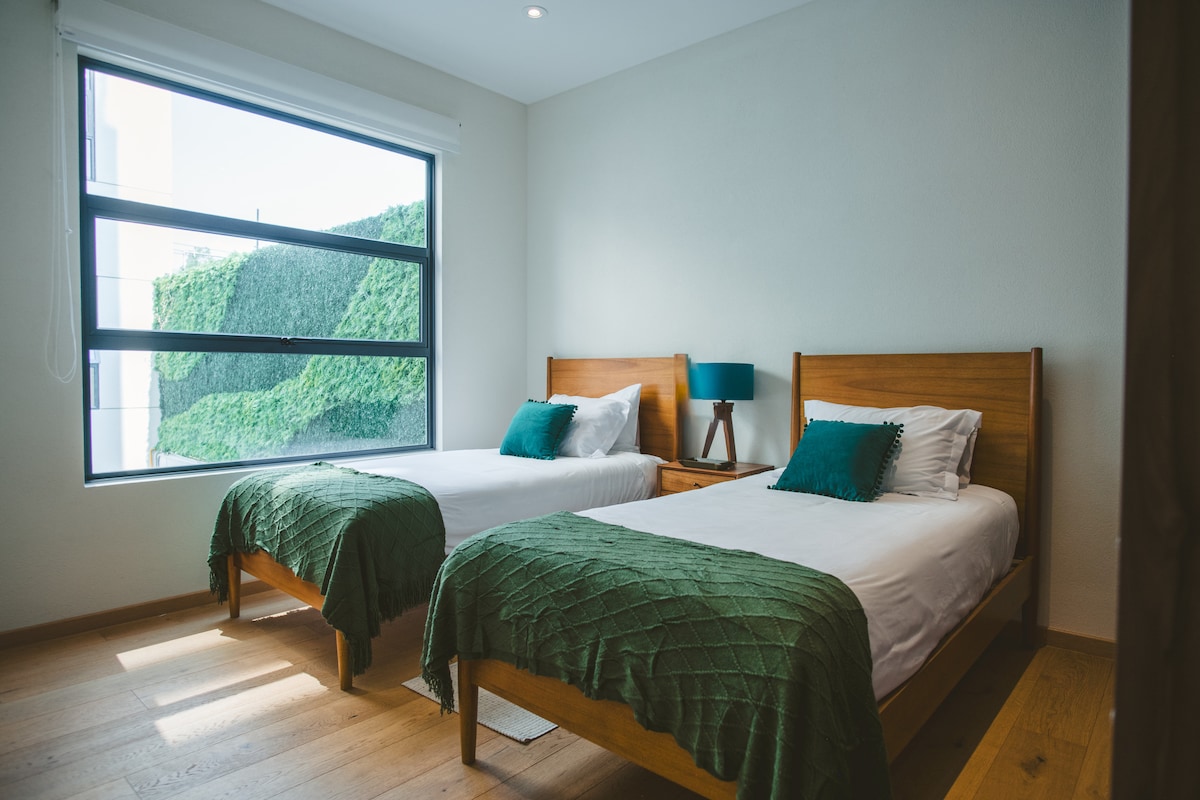 Two single beds are set within a sunlit room, adorned with green textured throws and decorative cushions. A bedside table with a blue lamp stands between the beds. A large window offers views of greenery outside, while light hardwood flooring enhances the spacious feel.