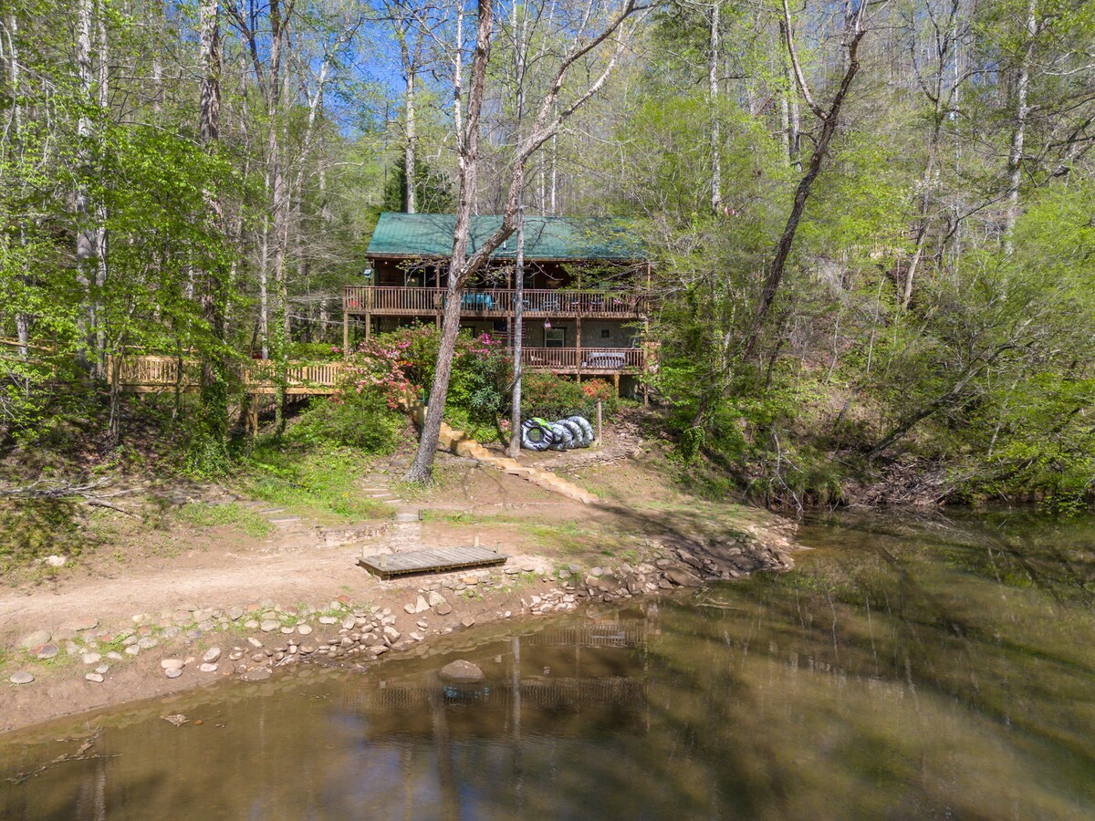 A log cabin retreat is nestled among trees, overlooking a calm river. A wraparound porch is visible, and outdoor seating is arranged on the deck. Kayaks and tubes are stored nearby, with natural landscaping enhancing the peaceful setting.