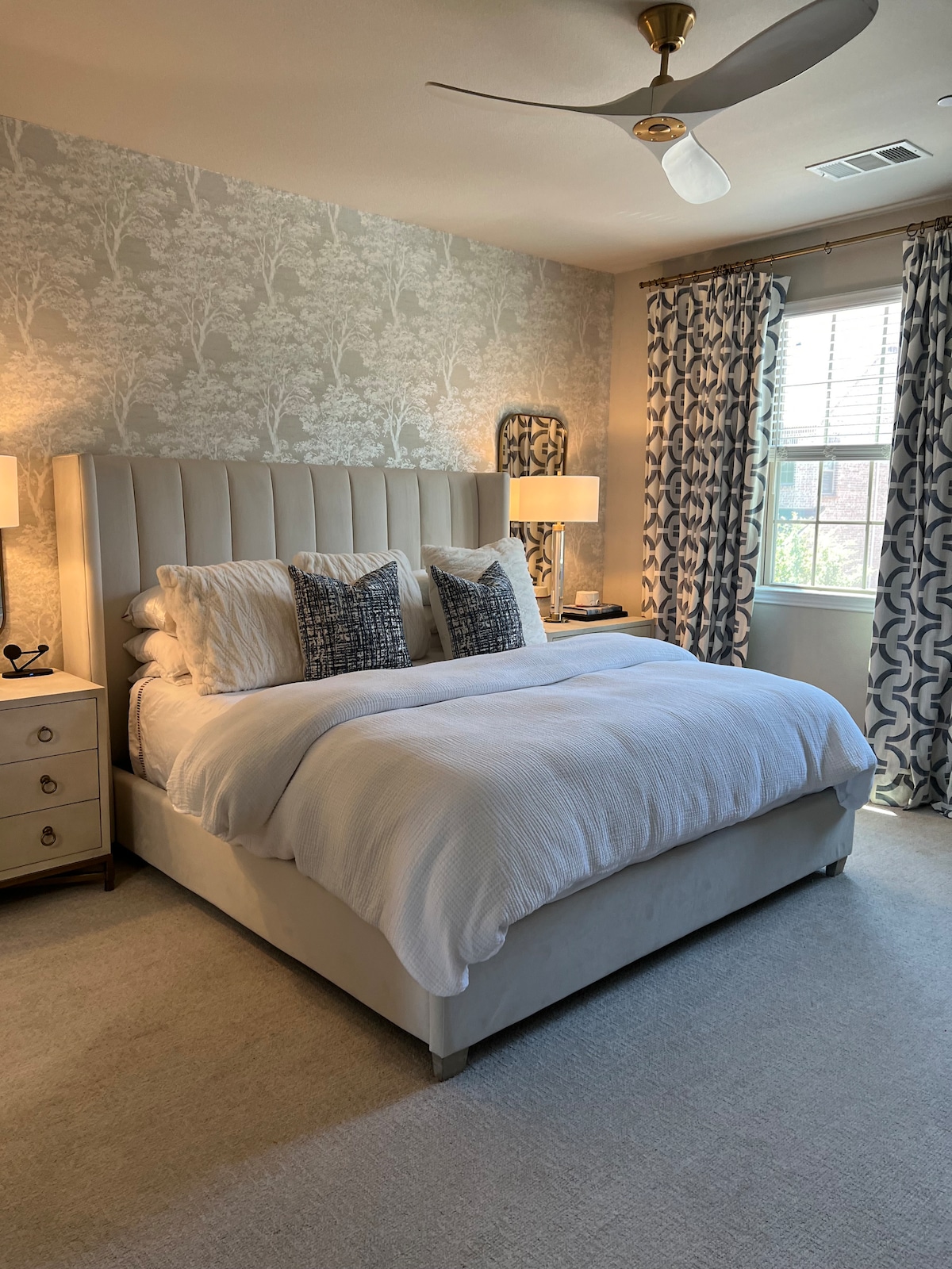 A spacious bedroom features a large, upholstered bed with decorative pillows. Nightstands with lamps are positioned on either side. Soft curtains frame a window, allowing natural light to illuminate the room. Subtle wallpaper adds texture to the walls, enhancing the overall aesthetic.