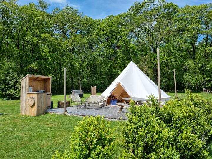 A spacious glamping tent is nestled within a serene green field. The tent features a wooden deck with outdoor seating and a small kitchen area nearby. Lush trees provide a natural backdrop, enhancing the peaceful setting. A wood-fired hot tub is also visible beside the tent.