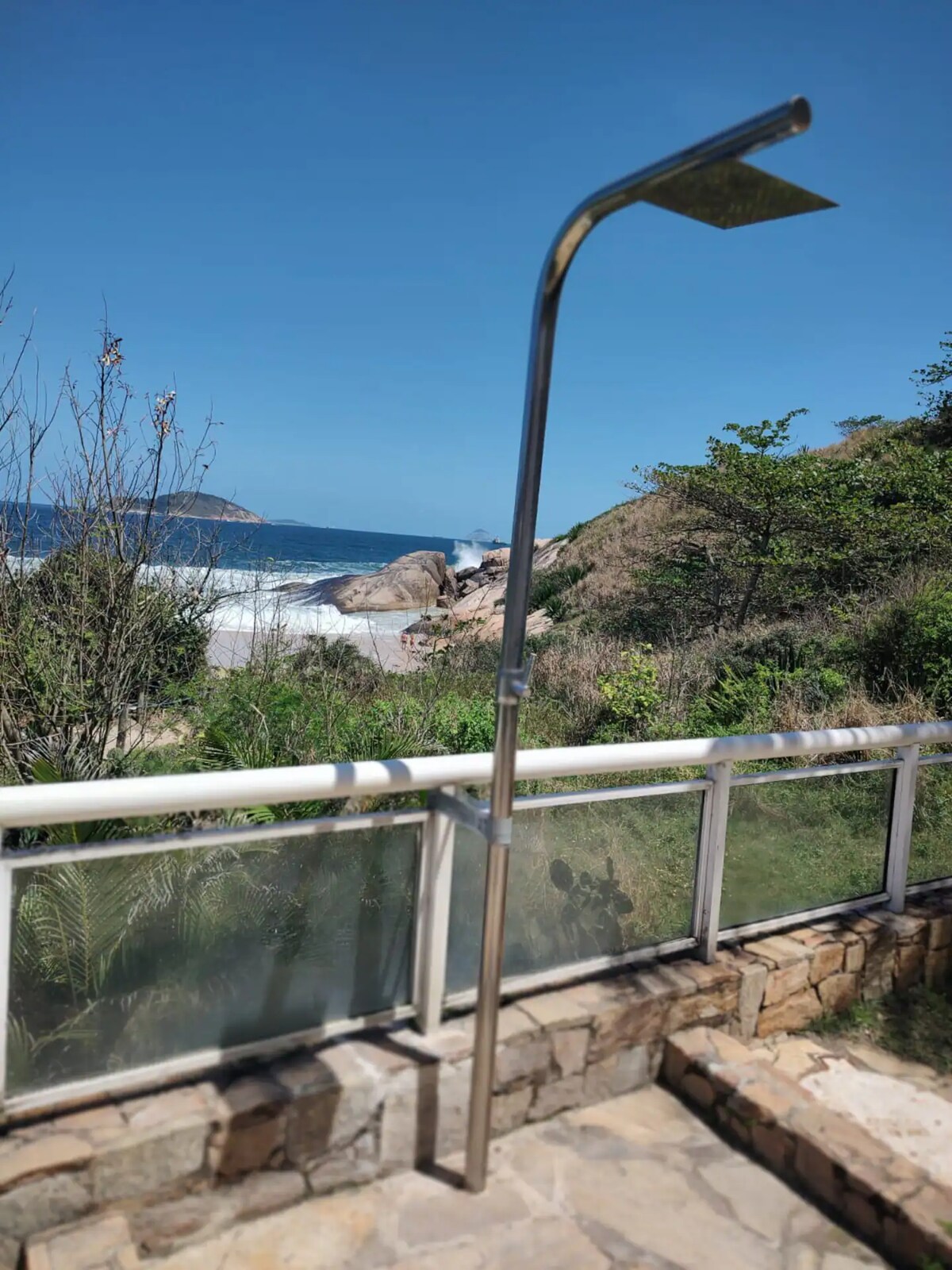An outdoor shower is positioned on a terrace, providing views of the beach and ocean. Lush greenery surrounds the area, and rocky formations are visible in the distance under a clear blue sky. The shower features a stainless steel design, emphasizing a modern aesthetic.