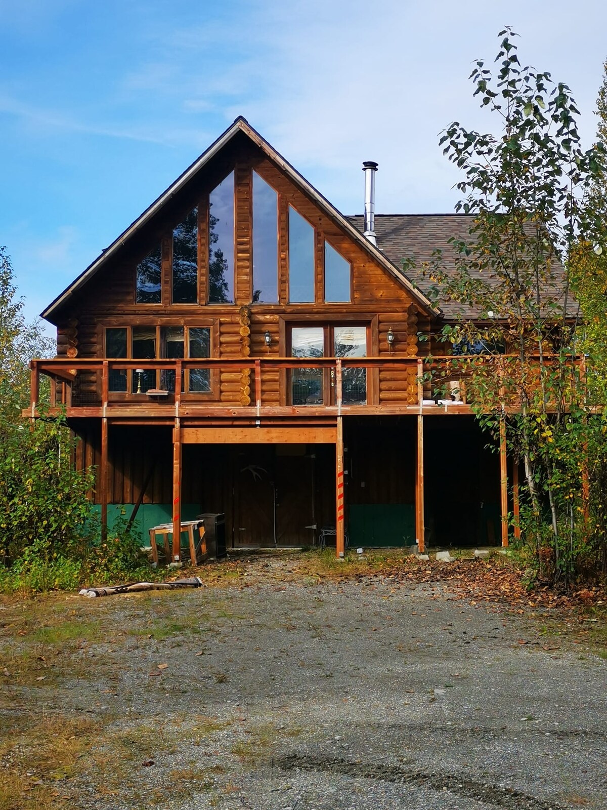 The cabin's exterior features a combination of large windows and rustic wood construction, surrounded by natural greenery. The spacious front porch is supported by wooden beams, and a gravel path leads to the entrance, inviting guests to explore the tranquil surroundings.