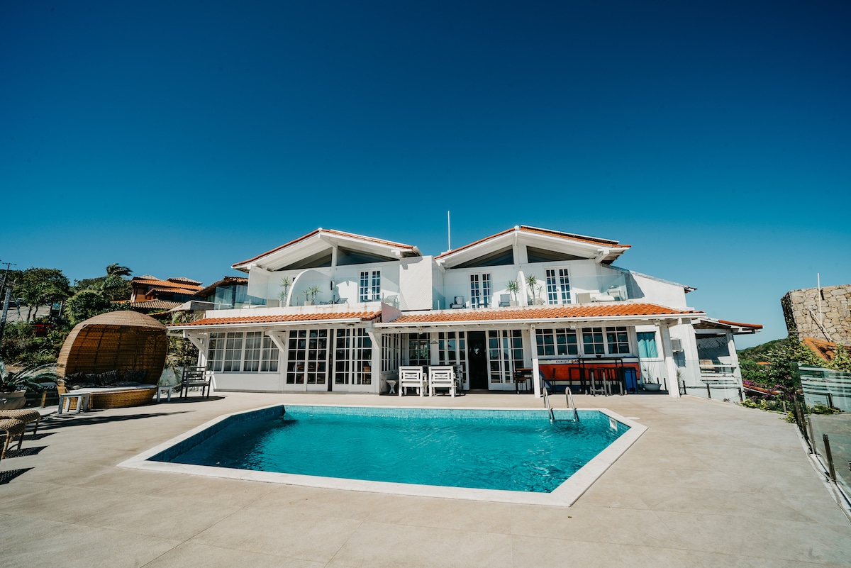 A spacious outdoor area features a swimming pool surrounded by a sunbathing deck. The two-story house, characterized by large windows and a sloped roof, faces the pool. Bright blue skies enhance the inviting atmosphere, creating a serene environment for relaxation.
