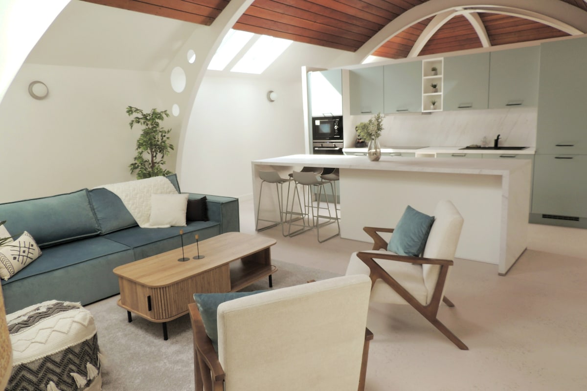 An open living area features a spacious blue sofa alongside two wooden armchairs, complemented by a wooden coffee table. The kitchen, visible in the background, includes a large island with bar seating, sleek cabinetry, and ample natural light from the skylights.