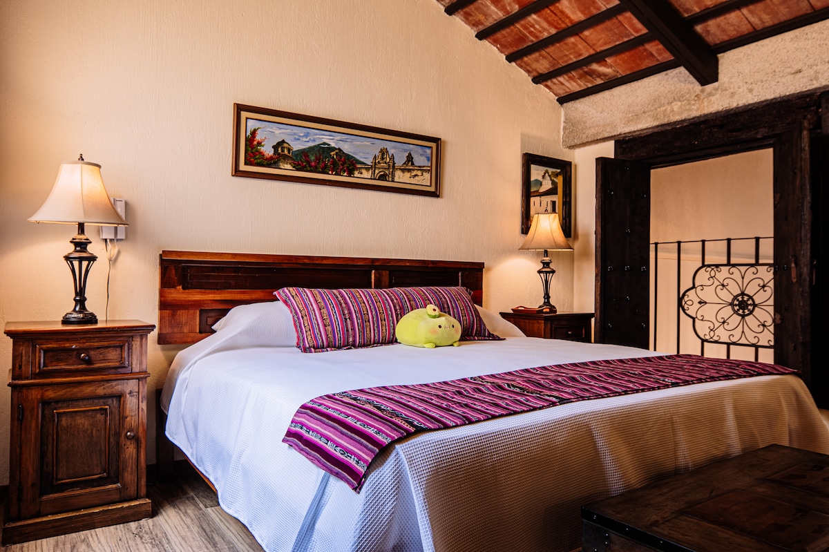 A spacious bedroom features a large bed adorned with a colorful woven blanket. Two bedside tables with lamps are positioned on either side of the bed. A decorative wall painting adds visual interest, while a wrought iron balcony railing is visible in the background.