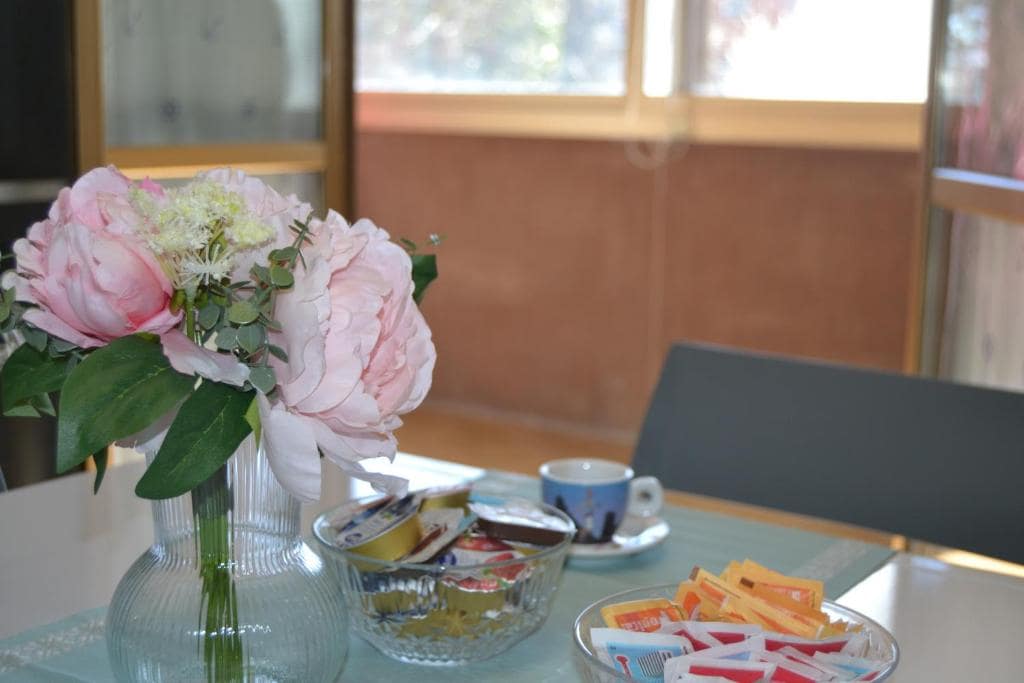 A dining table is adorned with a vase of large, light pink peonies and a bowl containing various tea bags and sweeteners. A small cup sits nearby, complemented by natural light streaming through large windows, creating a bright and welcoming atmosphere.