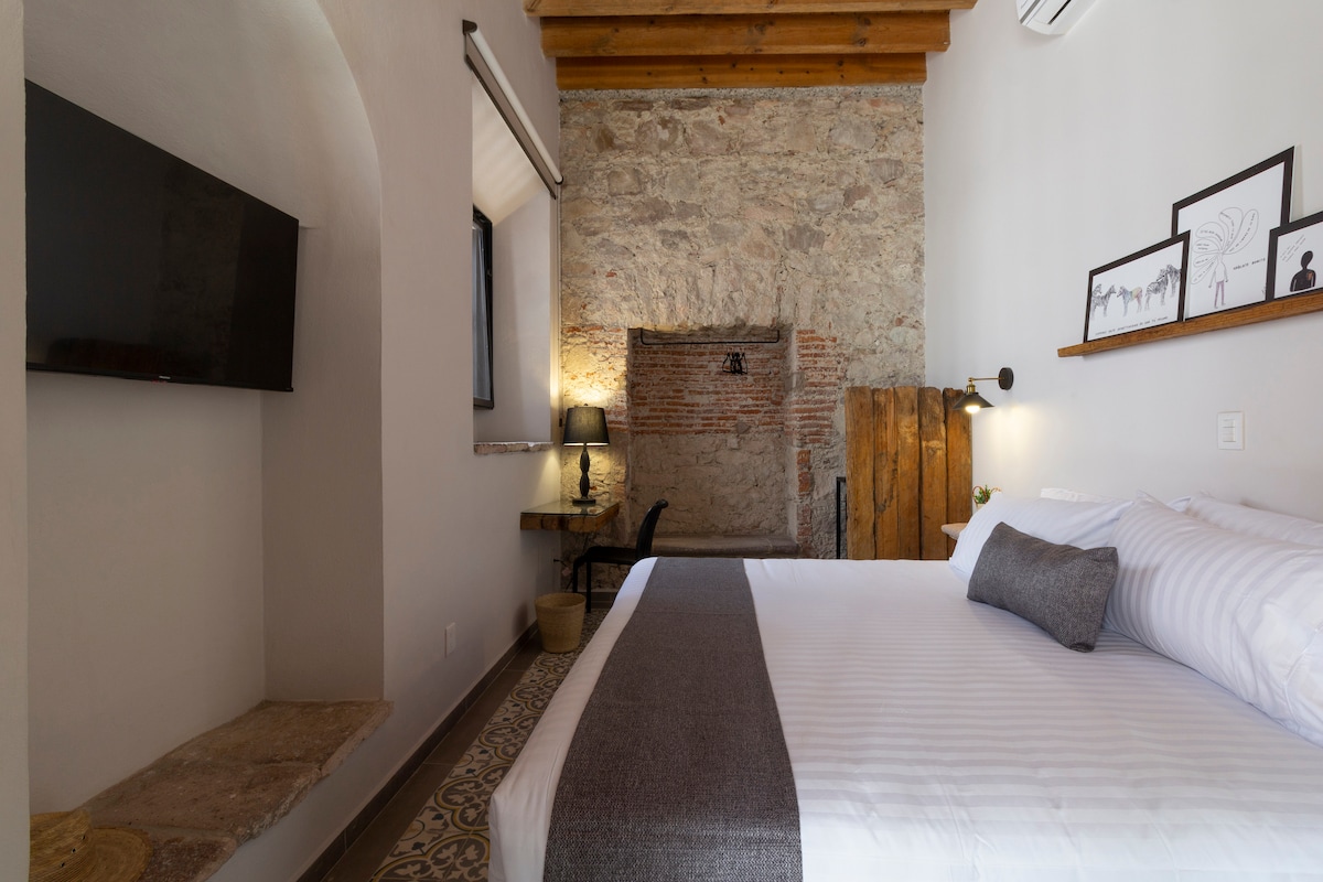 A spacious room features a large bed with subtle striped bedding and a decorative pillow. A flat-screen television is mounted on the wall. The room showcases exposed stone walls and wooden beams, with a wooden desk and chair positioned near a window.
