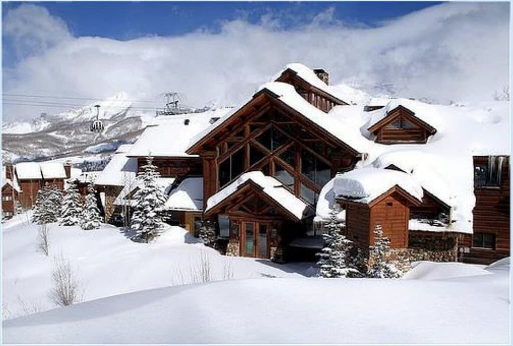 A rustic mountain lodge is nestled in a snowy landscape, showcasing wooden architecture and large windows that allow natural light inside. Snow-covered trees surround the property, enhancing its natural setting. The distant mountains are partially obscured by clouds, adding a serene backdrop.