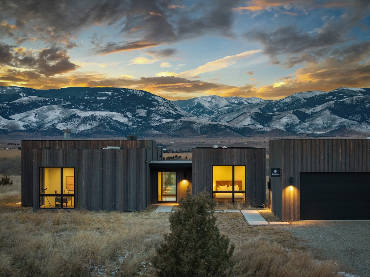 A modern home with a wooden exterior is set against a backdrop of mountains and a colorful sky. The two-section building features large windows illuminated from within, with surrounding grassy land and distant snow-capped peaks visible.