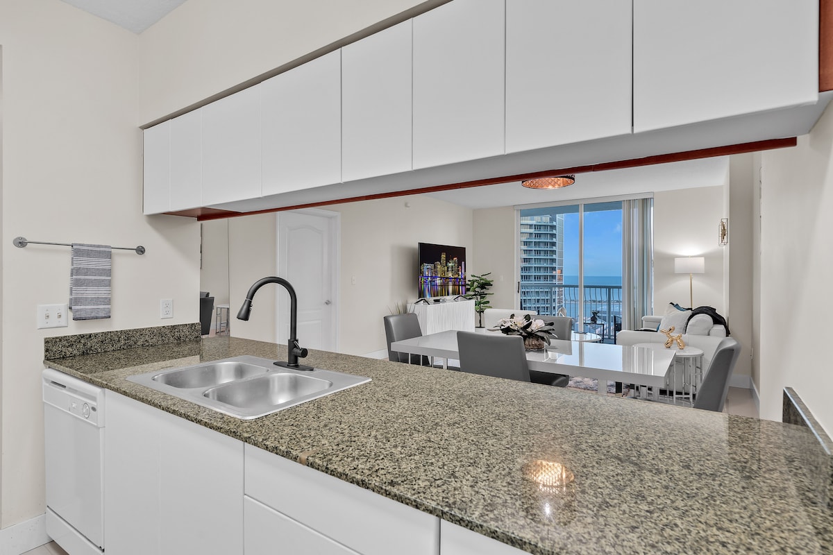 A modern kitchen showcases sleek white cabinetry paired with a speckled granite countertop. The space features a double sink and a dishwasher. A dining area is visible in the background, highlighted by a round light fixture and a large window offering views of the city.